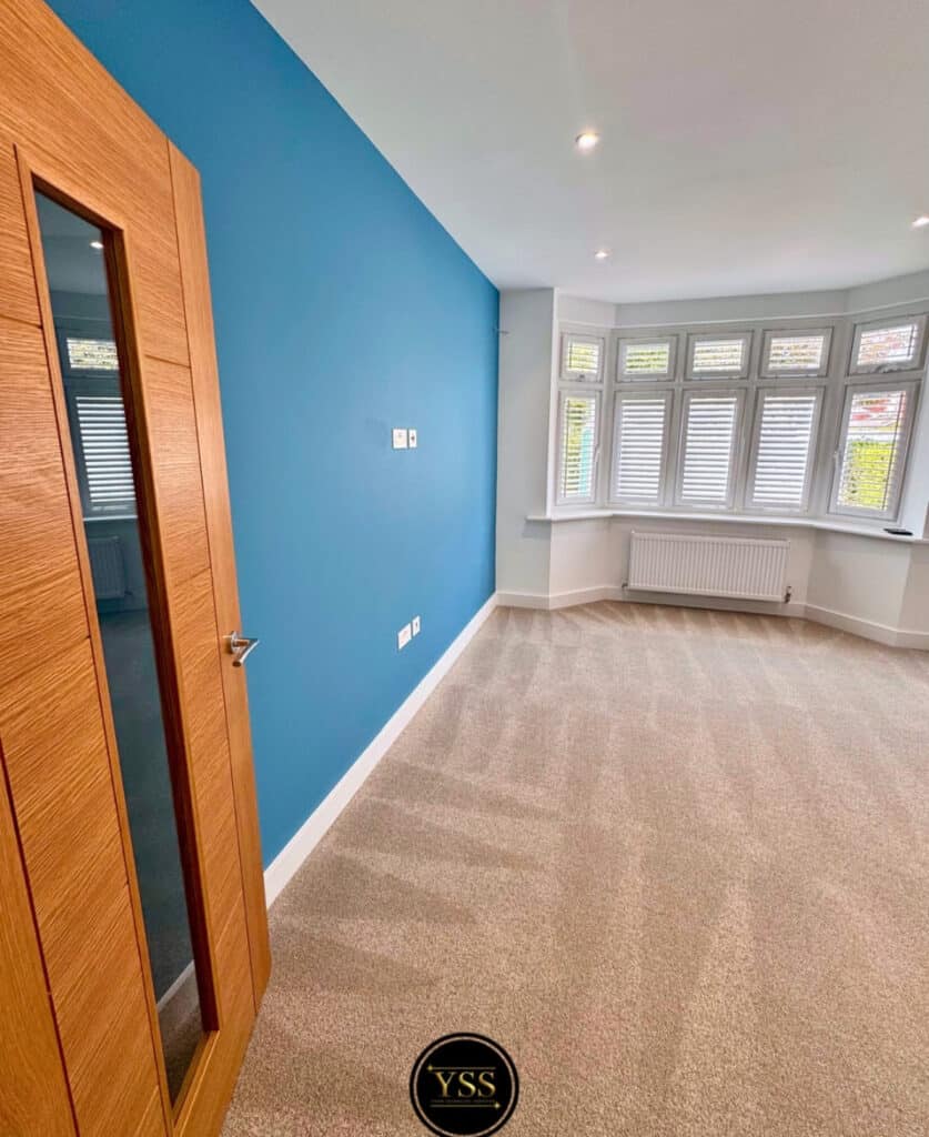 Bright, freshly decorated room with blue feature wall, large bay window, and beige carpet flooring.